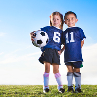 Ein Mädchen mit einem Fußball unterm Arm und ein Junge stehen im dunkelblauen Fußballtrikot Arm in Arm auf einer grünen Wiese.